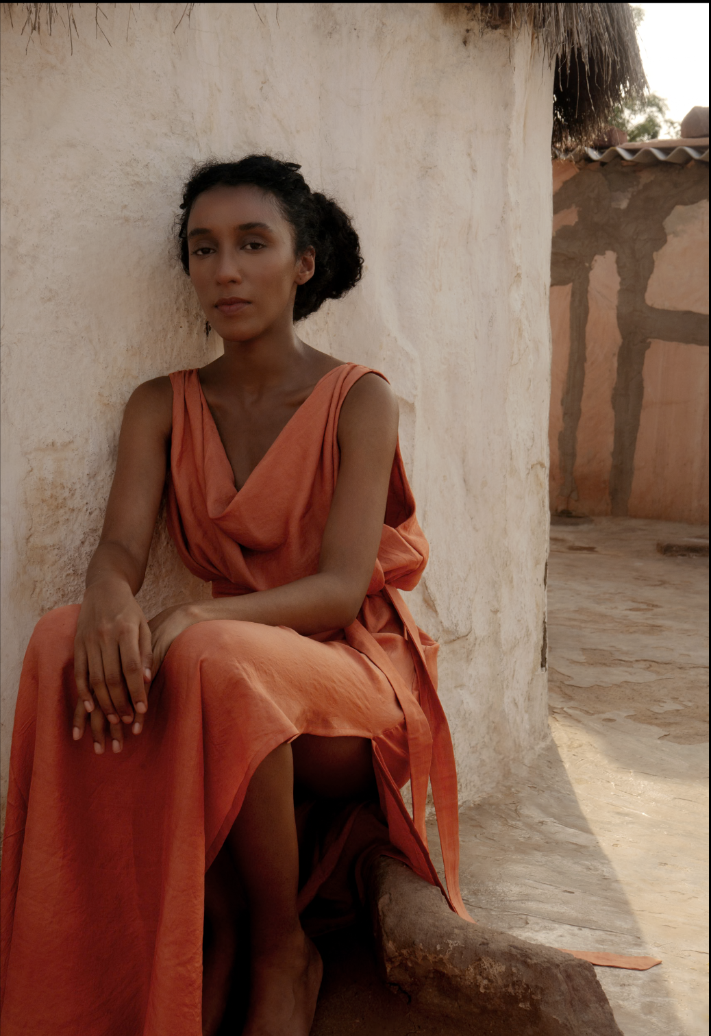 Hera dress in terracotta silk seated against a whitewashed wall